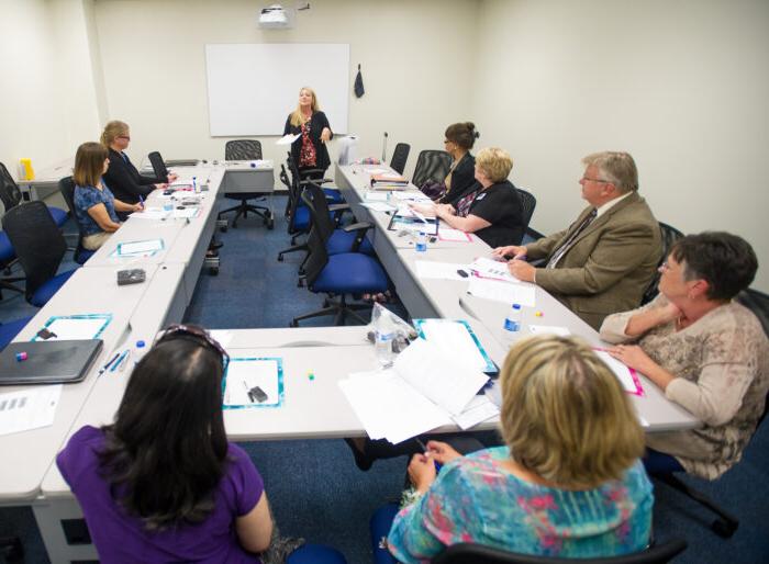 A group of professors sitting down at a table listening to one professor standing in front of them.