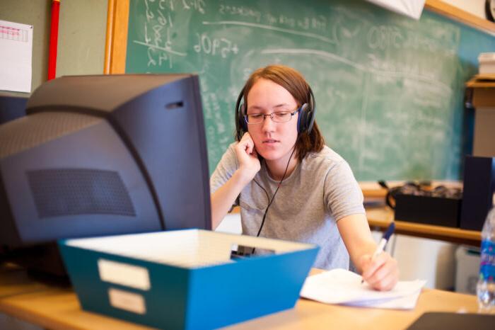 A young woman writing on paper while listing to headphones at a computer.