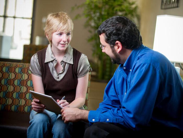 A student sits with a professor, attentively listening and taking notes during an informal one-on-one meeting.