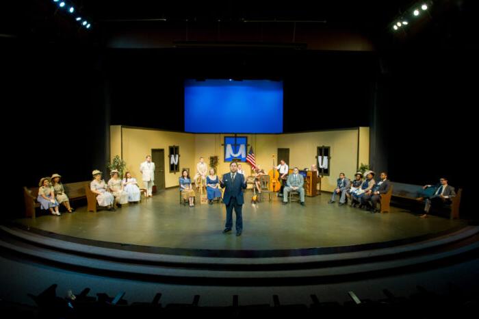 Lead actor standing on stage front with actors behind.