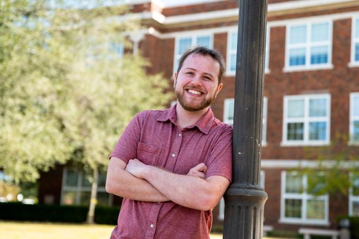 A smiling man in a short-sleeved red shirt stands outdoors, leaning against a black lamppost with arms crossed. A brick building with large windows is in the background, along with trees and greenery.