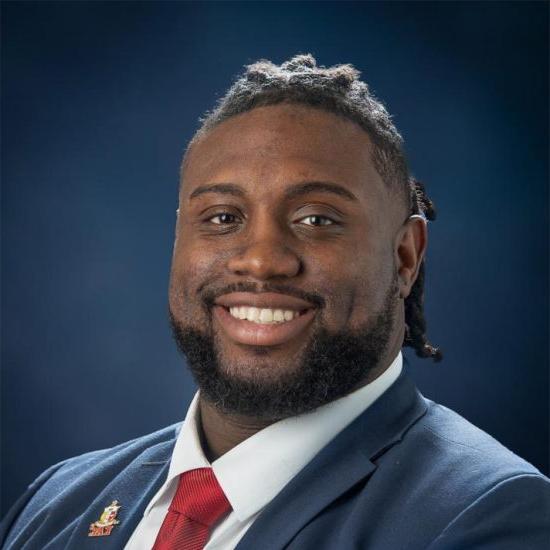 Portrait of a smiling man with a short hair and a beard, wearing a white shirt and blue blazer, is set against a dark blue background.