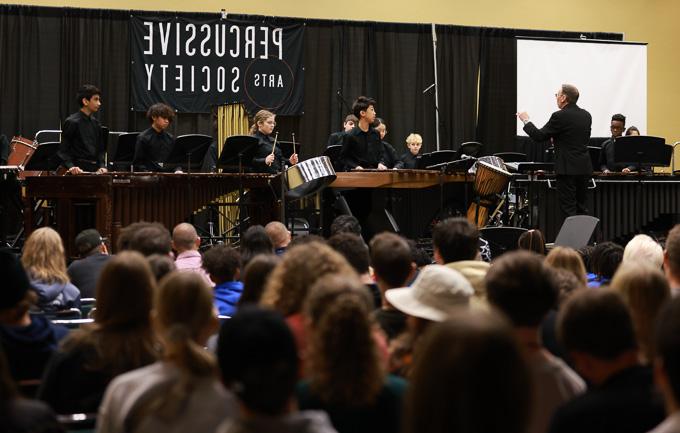 A group of musicians performs on stage with various percussive instruments, including a steel drum and marimbas, in front of an audience. A conductor in a black suit stands to the left, leading the ensemble. A "Percussive Arts Society" banner hangs in the background.