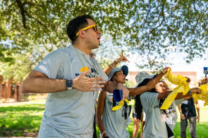 A group of enthusiastic students at an outdoor event cheer on their peers, led by a participant wearing sunglasses and holding a bandana, creating an energetic and spirited atmosphere.