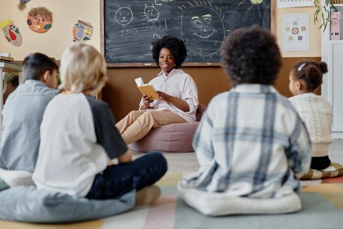 Teacher sits at the front of a classroom reading a book to several students who are sitting on cushions.