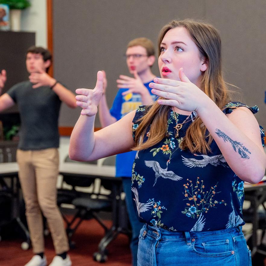 A young woman in a floral shirt gestures with her hands, focusing intently during a group activity, as others follow her movements.