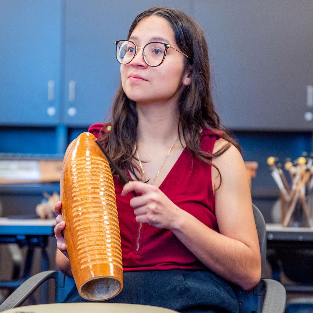 A young female wearing glasses and a red sleeveless shirt is looking forward and holding an instrument. The background appears to be in a classroom.